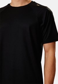 Sort atletisk t-shirt med ribbet tekstur, korte ærmer og accentdetaljer på skulderen. Materialet virker let og åndbart.