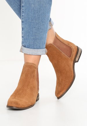 Bottines en daim marron avec panneaux latéraux élastiques et petit talon empilé, associées à un jean bleu clair retroussé.