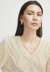 Pull en tricot beige avec un col en V, des motifs en relief de câbles et des manches en tricot ajouré. Des boucles d'oreilles en argent viennent accentuer le look.
