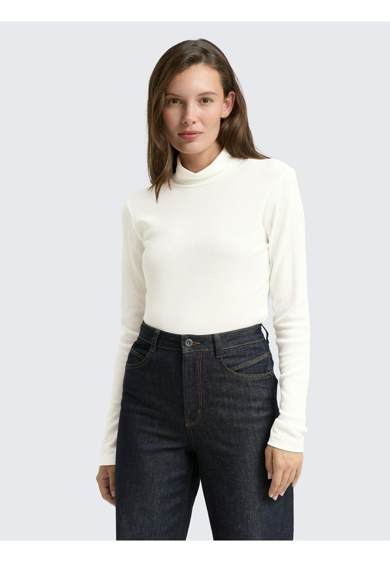 Femme aux cheveux bruns mi-longs portant un top blanc à manches longues et un pantalon en jean taille haute bleu foncé, se tenant devant un fond uni.