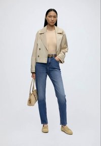 Femme portant une veste courte beige, un top beige, un jean bleu taille haute, des mocassins beiges, tenant un sac à main beige assorti sur fond uni.