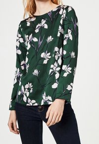 Grön blommig blus med långa ärmar, med vita och lila blommönster mot en texturerad bakgrund. Rundad halsringning och figurnära design.