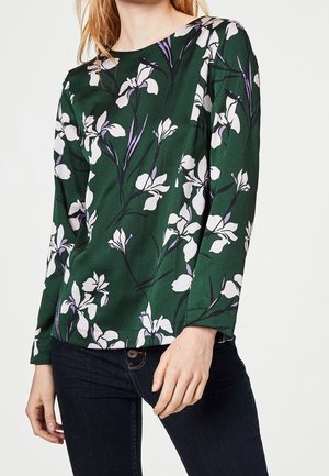 Blouse verte à fleurs avec manches longues, présentant des motifs de fleurs blanches et violettes sur un fond texturé. Encolure arrondie et coupe ajustée.