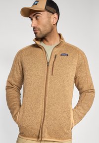 Brūna flīsa jaka ar rāvējslēdzēju, teksturētu virsmu un sānu kabatām. Ir augsts apkaklis un neliels logotips krūšu rajonā.