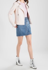 Helle rosa Kunstlederjacke mit silbernen Reißverschlüssen, kombiniert mit einem Jeansrock in Blau und metallischen, silbernen Ankle Boots. Glatte Texturen sind sichtbar.