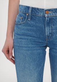 Gros plan sur une personne portant un jean en denim bleu taille haute et une chemise blanche rentrée, la main gauche détendue le long du corps.