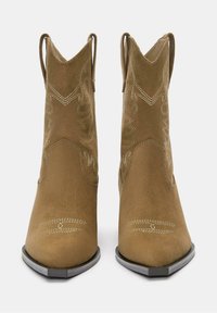 Bocanci de cowboy din suede maro, cu vârfuri ascuțite și broderie în stil vestic. Au prinderi scurte pe laterale și o talpă texturată de culoare gri.