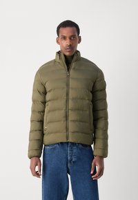 Olivengrøn pufferjakke med vandret quiltning, høj krave, fuld lynlås foran og logo accent nederst til venstre. Stylet med blå jeans.