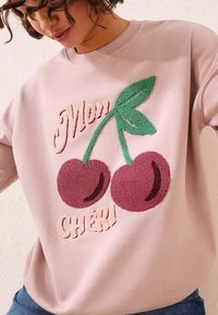 Felpa rosa chiaro con ricamo testurizzato di ciliegie e foglie verdi, con la scritta "Mon Chéri." Taglio casual con collo rotondo.