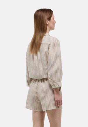 Femme aux cheveux lisses châtain clair portant une chemise à manches longues beige et blanche à rayures verticales et un short assorti, vue de profil.