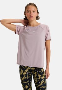 Woman wearing a lavender short-sleeve sports shirt and black leggings with yellow and pink abstract floral patterns, posing with one hand behind neck.