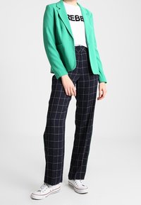 Veste verte portée sur un t-shirt blanc avec le texte noir "REBEL", associée à un pantalon à carreaux bleu marine et des baskets blanches.