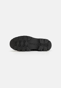 Semelle de chaussure en caoutchouc noir avec des motifs de crans profonds et des stries prononcées. Logo de la marque estampé visible. Surface texturée pour une meilleure adhérence.