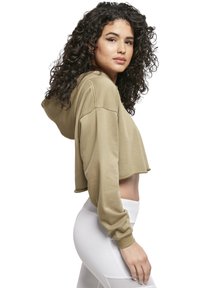 Beige színű, pamut cropped kapucnis pulóver, hosszú ujjakkal, elülső kenguruzsebbel és laza szabással. A modellen fehér, magas derekú leggings van.