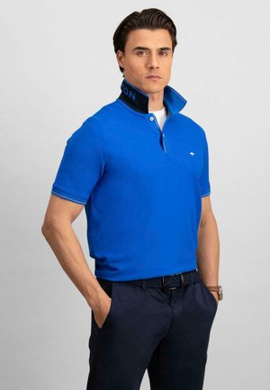 Blå polo t-shirt med opretstående krave med sorte og blå detaljer, båret med mørke bukser. Materialet ser glat og tætsiddende ud.