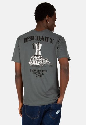 Mann trägt graues T-Shirt mit schwarzem Text "IRIEDAILY" und "SLOWPRESSO COFFEE CLUB" sowie einer Grafik einer Schildkröte, die eine Kaffeemaschine trägt.