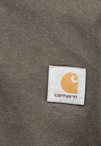 Λευκή τετράγωνη υφασμάτινη ετικέτα με πορτοκαλί λογότυπο σε σχήμα ημισελήνου και κεί μενο "carhartt" σε φόντο από χονδρό, καφέ υφαντό ύφασμα.