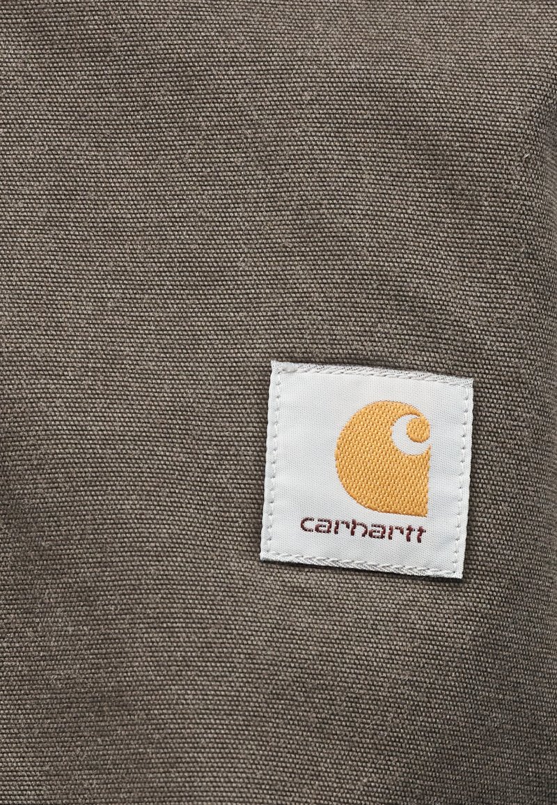 Weißes quadratisches Stoffetikett mit einem orangen Mondsichel-Logo und dem Text "carhartt" auf grobem braunem Gewebehintergrund.