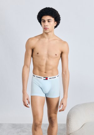 Boxer en coton bleu clair avec une ceinture blanche affichant le logo « TOMMY HILFIGER ». Le modèle est debout dans une pose neutre, les bras détendus.