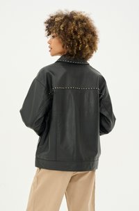 Melna mākslīgās ādas jaka ar brīvu piegriezumu, sudraba kniedes detaļas ap apkakli un muguru, kas kontrastē ar beigu biksēm.