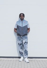 Sivi prsluk na napuhavanje s uzorkom quilted dizajnom preko svijetlosivog majice, spojeno sa sivim duksericama u kamuflažnom uzorku i bijelim tenisicama.