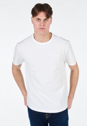T-shirt bianca in cotone con colletto rotondo e maniche corte, caratterizzata da un logo discreto sul lato sinistro. Indossata con jeans blu.