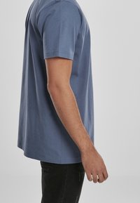 Boční pohled na osobu oblečenou v modré košili s krátkým rukávem a černých kalhotách na jednobarevném světlém pozadí.