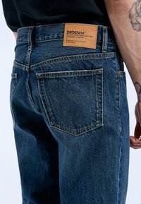 Jeans de mezclilla azul oscuro con una etiqueta de cuero marrón claro etiquetada como "DRDENIM" en la cintura trasera, con un diseño clásico de cinco bolsillos y costuras cosidas.
