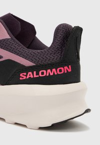 Κοντινό πλάνο φτέρνας μοβ και μαύρου αθλη τικού παπουτσιού Salomon με ροζ λογότυπο και παχιά λευκή σόλα σε λευκό φόντο.