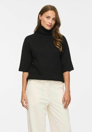 Femme aux longs cheveux portant un pull à col roulé noir avec des manches jusqu'au coude et un pantalon clair taille haute sur fond blanc.