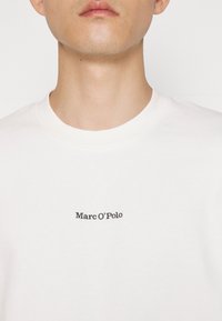 Gros plan sur une personne portant un t-shirt blanc à col rond avec « Marc O'Polo » imprimé en petites lettres noires sur la poitrine.