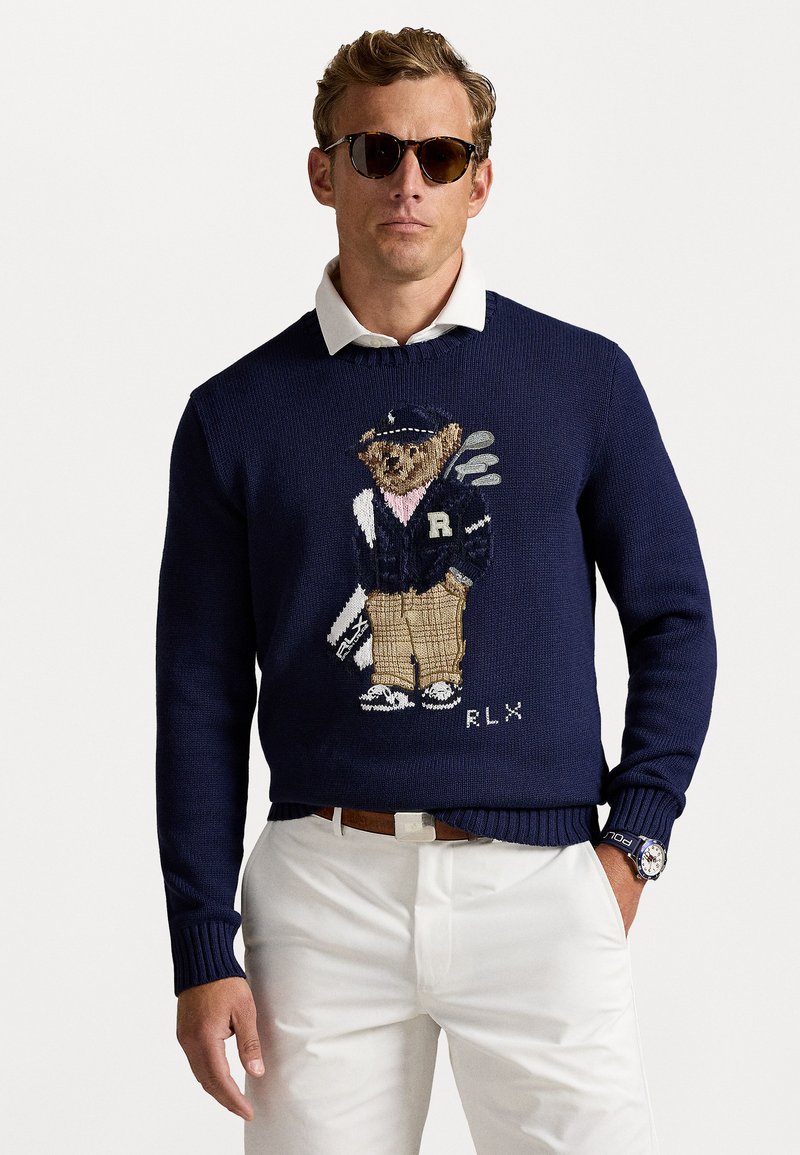 Maglione lavorato a maglia blu navy con grafica di un orso vestito da golf, con pantaloni beige e giacca blu, abbinato a pantaloni bianchi.