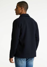Navyblauwe jas van gestructureerde stof, met een kraag en zijsplitten. Heeft een knoopsluiting bij de manchetten en stiksels op de achterkant.