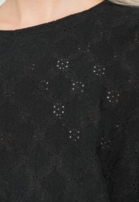 Μαύρο ύφασμα με υφή σε διαμαντένιο σχέδιο και μικρές ανθοδέσμες, φορεμένο κοντά στην περιοχή του λαιμού και του ώμου.