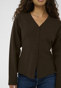 Tumši brūns adīts kardigans ar V veida kakla izgriezumu, garām piedurknēm un septiņām priekšējām pogām. Dizains ietver nedaudz izliektu apakšmalu un maigu tekstūru.