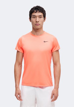 Mann trägt ein hellorangefarbenes Nike-Kurzarmshirt und weiße Shorts, steht frontal vor einem schlichten hellen Hintergrund.