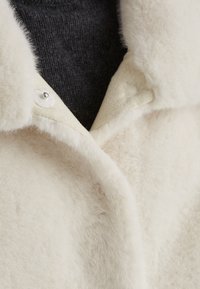 Cremfarbener Mantel mit einer weichen, flauschigen Textur. Verfügt über einen Reverskragen und einen Druckknopfverschluss, kombiniert mit einem dunkelgrauen Strickoberteil darunter.