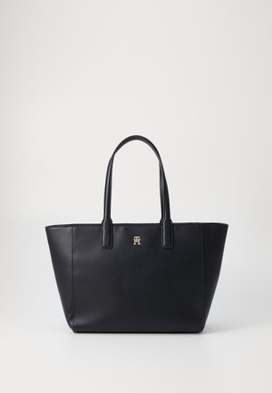 Schwarze Leder-Tote-Tasche mit strukturiertem Design, zwei langen Griffen und einem metallischen Logoakzent auf der Vorderseite. Glatte Textur, minimalistischer Stil.