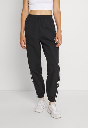 Osoba nosząca czarne spodnie dresowe Adidas z białymi paskami, białe trampki i biały krótki top, stojąca na jednolitym tle.