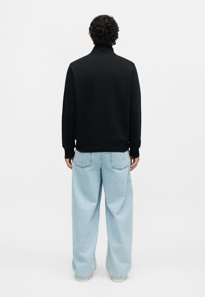 Άτομο με σγουρά σκούρα μαλλιά που φοράει μαύρο μακρυμάνικο τοπ και ανοιχτό μπλε φαρδιά τζιν, στραμμένο με την πλάτη σε απλό φόντο.