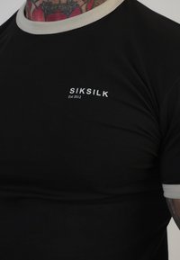 Črna majica s prilagojeno zasnovo, ki ima svetlo sivo rebrasto okroglo ovratnik in majhen beli logotip s "SIKSILK" in "Est 2012" na prsih.