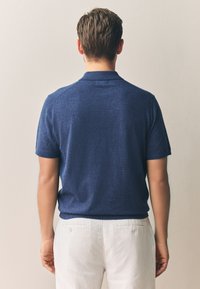 Marineblaues Polohemd aus strukturiertem Stoff, mit klassischem Kragen, kurzen Ärmeln und gerippten Bündchen. Kombiniert mit hellfarbenen Hosen.