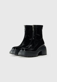 Black patent leather ankle boots with front zippers and chunky soles with deep treads on a plain light background.