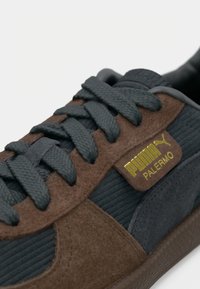 Sneakers ar tumši pelēku tekstilmateriālu un brūnu zamšu, ar strukturētām svītrām, zaļām auklām un zelta logo uzlīmi.