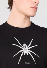 Černé bavlněné tričko s velkým bílým potiskem pavouka ve středu a slovem "BOSS" níže v jemném písmu.
