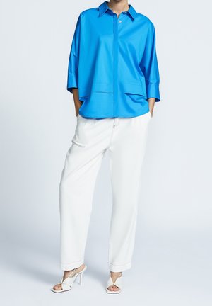 Donna che indossa una camicia blu brillante e larga, pantaloni bianchi a gamba larga e sandali bianchi con tacco alto, in piedi con le mani nelle tasche.