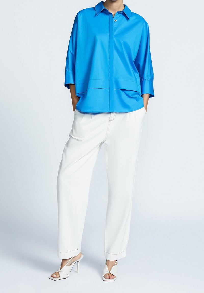Femme portant une chemise ample bleu vif, un pantalon blanc à jambes larges et des sandales blanches à talons hauts, debout les mains dans les poches.