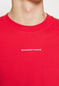 T-shirt di cotone rosso con collo rotondo; presenta una ricamo bianco con la scritta "SANDRO PARIS" centrata sul petto. Texture liscia.