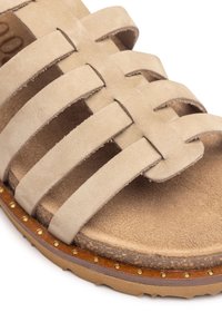 Beige Wildleder-Sandale mit mehreren geflochtenen Riemen und einem strukturierten Fußbett, mit einer braunen Sohle, die mit kleinen goldenen Nieten akzentuiert ist.