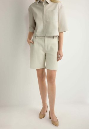 Kobieta ubrana w beżową koszulę z krótkim rękawem z kołnierzykiem oraz pasujące beżowe krótkie spodenki do kolan, stojąca na tle jednolitego tła, w beżowych butach na blokowym obcasie.